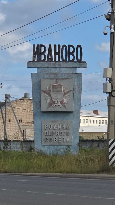A tall, light blue monument with Cyrillic text is positioned in the foreground against a bright blue sky with scattered clouds. The monument features a large emblem with a red star, a hammer and sickle, and a laurel wreath. Below the emblem, the words "РОДИНА ПЕРВОГО СОВЕТА" (Motherland of the First Soviet) are inscribed in raised lettering. The monument is flanked by a chain-link fence with barbed wire and overgrown grass. In the background, to the left, is a brick building with a visible ventilation pipe. To the right, a utility pole with surveillance cameras and electrical equipment stands tall. A road with white dashed lines runs across the bottom of the frame. This monument is located in Novo-Talitsy, Ivanovo Oblast, Russia.