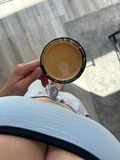 A first-person perspective shot shows a hand holding a glass of coffee. The hand is fair-skinned, and the coffee glass has a dark rim. The coffee itself is light brown with a foamy top. The person is wearing a light blue top with a dark trim, and part of light-colored drawstring shorts are visible. The background consists of light grey flooring and a grey rug with a shadow cast upon it. The location cannot be determined from the image.
