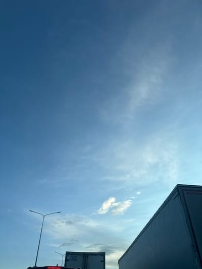 The image depicts a clear blue sky with scattered wispy clouds. A slender crescent moon is visible in the upper left quadrant of the sky. In the foreground, the top portions of two large trailer trucks are visible, their gray sides extending upwards and across the frame. The truck on the right has red lettering on its side that reads "Kässbohrer". A street lamp with two lights is also visible on the left side of the frame. The scene appears to be on a road, possibly a highway, given the presence of the trucks and street lamp. The location is identified as Krutitsy, Russia.
