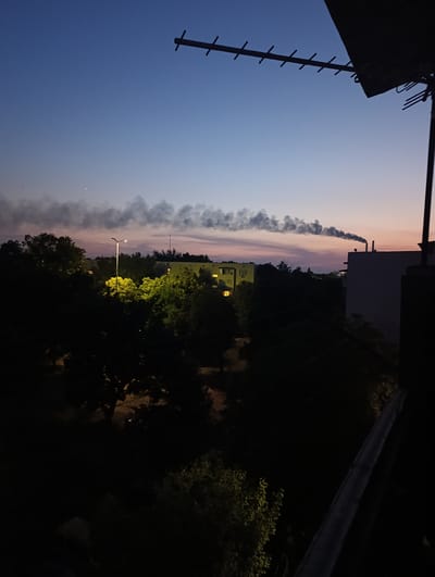 In Ladyzhyn, Ukraine, the image captures a twilight scene with a clear sky transitioning from deep blue to soft pink. A prominent antenna is visible in the upper right, silhouetted against the sky. In the middle ground, smoke billows from a chimney, creating a dark plume against the colorful horizon. Buildings and trees are visible in the cityscape, with some windows illuminated by artificial light. A streetlamp in the mid-left foreground casts a bright yellow glow on the surrounding trees. The overall impression is of a quiet evening in a residential area.