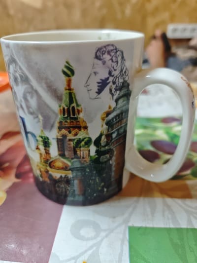 A white ceramic mug with a handle is positioned on a patterned tablecloth. The mug is decorated with an illustration featuring a profile of a man with curly hair and a stylized depiction of a Russian orthodox cathedral. The background appears to be a blurred interior. The tablecloth has a design with floral elements. The image is taken from a medium distance, with the mug being the primary subject. The scene is indoors, and there is no indication of the city of Konokovo, Russia.
