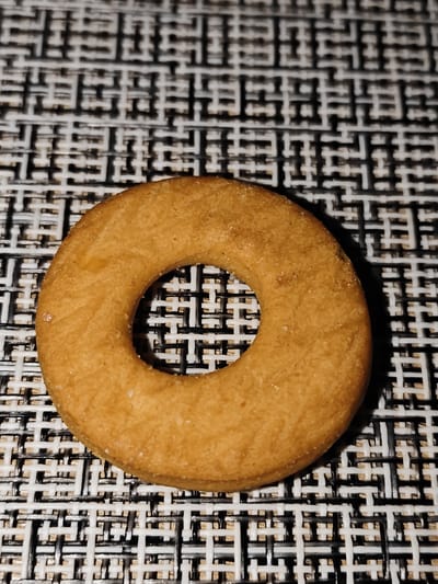 A single, golden-brown, ring-shaped cookie rests on a black and white patterned placemat. The cookie has a slightly rough texture and appears to be made of a dense dough, possibly shortbread or a similar biscuit. Its surface exhibits subtle variations in color, with some areas appearing lighter and others slightly darker, suggesting it has been baked evenly. A few small, granular specks, possibly sugar crystals or crumbs, are visible on the cookie's surface. The placemat beneath the cookie features an intricate geometric pattern formed by interwoven black and white straps, creating a criss-cross effect. The lighting is direct and appears to be from above, casting a soft shadow from the cookie onto the placemat. The image is a close-up shot, focusing primarily on the cookie and its immediate surroundings. There are no people or any other objects present in the frame. This image does not contain any information related to the city of Bugulma, Russia.