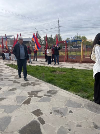A group of people holding Armenian flags are walking on a stone-paved path in Akhuryan, Armenia. One man is wearing a hat and jacket, while the others are dressed in casual clothing. A playground is visible in the background with swings and other play equipment. Overhead are electrical wires. The sky is overcast.