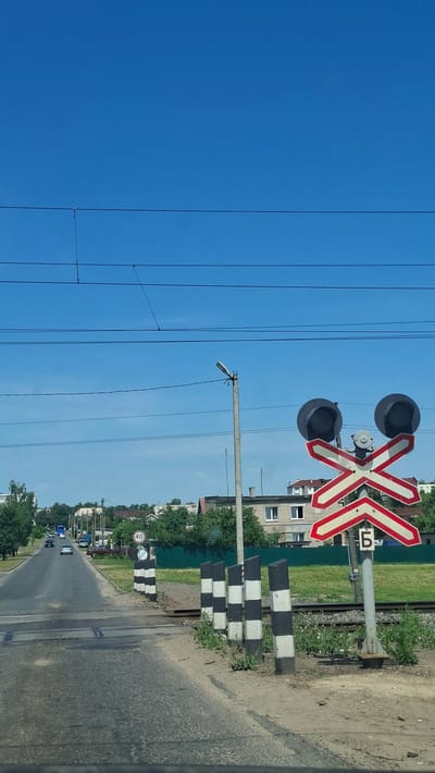 The image depicts a railway crossing on a clear, sunny day in Dzyarzhynsk, Belarus. A paved road intersects a set of railroad tracks, with a railway signal displaying two black, circular lights on a red and white crossbuck sign. The signal is mounted on a gray pole that also has a Cyrillic letter "Б" on it. Adjacent to the road are several black and white striped posts, and a speed limit sign indicating "40" is visible further down the road on the left. In the background, there are residential buildings and trees. A few cars are present on the road, and overhead power lines are visible against the bright blue sky.