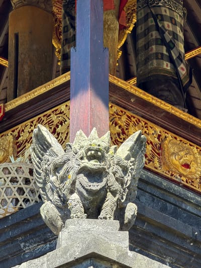 The image shows an ornate stone carving of a mythical creature, possibly a dragon or a demon, integrated into the architecture of a temple or traditional building. The carving is grey and textured, with intricate details on its face, including sharp teeth, wide eyes, and a headdress. It appears to be mounted on a dark, textured stone structure, possibly part of a pillar or wall.
Below and to the left of the carving, there is a large wooden beam, a portion of which is illuminated by sunlight. This beam is positioned diagonally across the frame. Behind the beam and above the carving, there are more architectural elements. A piece of dark fabric with a black and white checkered pattern, interspersed with small red, yellow, and green motifs, is draped over a wooden structure. This fabric is trimmed with a gold-colored decorative band. Further back, golden ornate carvings with floral motifs are visible against a red background, set within wooden frames. The overall setting suggests a place of cultural or religious significance, likely in a tropical or subtropical region given the presence of sunlight and the style of architecture and decoration. The time of day appears to be daytime, with strong sunlight casting shadows. There is no visible text in the image.