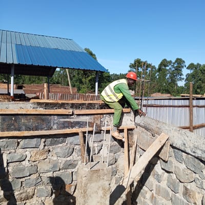 In Borabu Chitago ward, Kenya, a construction worker wearing a red hard hat, a fluorescent yellow safety vest over green clothing, and tan shoes is positioned atop a partially constructed stone wall. He appears to be inspecting or adjusting the framework. The wall consists of irregularly shaped grey stones. Wooden planks and concrete forms are used to shape the structure, with reinforcing steel bars visible. In the background, a blue and grey corrugated metal roof covers an open structure. Beyond that, trees and other buildings are seen under a clear blue sky.