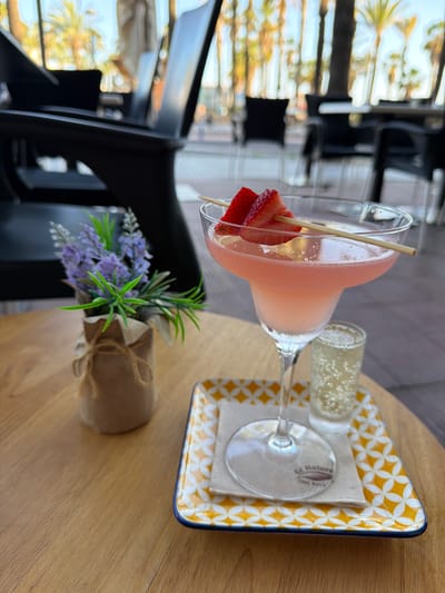 The image displays a close-up view of two refreshing drinks and a decorative plant arranged on a light wooden table, likely in an outdoor or semi-outdoor setting in Salou, Spain.

The main subject is a frosty, pinkish-red cocktail, possibly a frozen daiquiri or margarita, served in a classic martini glass. It is garnished with two slices of fresh strawberry impaled on a small wooden skewer, which rests across the rim of the glass. Adjacent to this, a smaller shot glass contains a clear, sparkling beverage, identifiable by the numerous tiny bubbles clinging to its side.

Both glasses are presented on a distinctive square ceramic tray featuring a bright yellow and white geometric pattern, with a dark blue rim. Beneath the martini glass, a white napkin is visible, bearing partial text that appears to read "GC Natura" and "100% Recy," likely indicating "100% Recycled."

To the left of the tray, a small decorative plant, resembling lavender with its purple flowers and green foliage, is placed on the table. It is wrapped in rustic brown paper and tied with twine, adding a touch of natural decor.

The background is softly blurred but suggests an inviting atmosphere. Distant elements include reflections of what appear to be palm trees and brightly lit areas, hinting at a sunny day. Modern, dark-colored furniture, possibly chairs or tables, can also be discerned in the blurred mid-ground, suggesting a cafe or bar environment. The overall scene conveys a sense of relaxation and leisure, typical of a vacation spot.