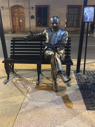 The image shows a bronze statue of a seated man, likely a historical figure, placed on a black park bench. The statue is positioned in a public space, with a building featuring barred windows and a large wooden door visible in the background. The statue is near a streetlight post with a poster that includes a portrait of a man and text, "BASADRE ENTRE NOSOTROS", suggesting it is related to Jorge Basadre Grohmann, a noted Peruvian historian from Tacna. The ground is paved with light-colored stone tiles. The location is indicated as Tacna, Peru.