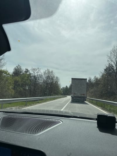 The image, rotated 90 degrees clockwise, presents a view from the interior of a car looking forward through the windshield. The perspective suggests the car is in motion, driving on a road or highway.
In the foreground, elements of the car's interior are visible. On the right side, a black dashcam is mounted on the windshield, casting a shadow on the dashboard. Below it, a speaker grille with a hexagonal mesh pattern is integrated into the dashboard. A small portion of the driver's side A-pillar is visible on the left.
The road stretches ahead into the distance, with white lines marking what appear to be lanes. A metallic guardrail runs along the right side of the road. Ahead, a large, rectangular vehicle, likely the rear of a truck or trailer, is visible in the distance. Lush green trees and bare-branched trees line both sides of the road, suggesting a rural or wooded area.
The sky overhead is bright but partially cloudy or overcast, indicating daytime conditions, possibly mid-morning or afternoon. A small, blurry yellow object or dot is faintly visible in the far distance, near the horizon, slightly to the left of the truck, though its exact nature is unclear.
The scene depicts a typical road trip or commute in a rural setting, with the vehicle following another on a relatively clear day. The location is specified as Dobrinovo, Bulgaria. No visible text is present in the image.