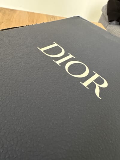 A close-up photograph depicts the upper corner of a grey Dior box with the word "DIOR" printed diagonally across its surface in a light-colored font. The box has a textured surface. In the upper left corner, there are blurred outlines of soft clothing and a wooden surface. A white wall is visible on the left side of the image. The photograph's location is unknown.