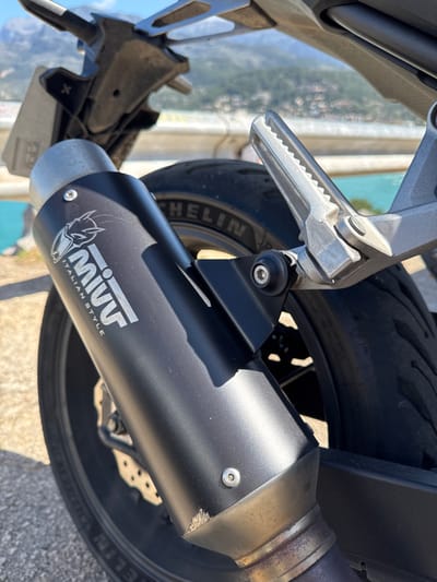 The image provides a forensic close-up of the rear section of a motorcycle, parked at a viewpoint overlooking a coastal area in Sóller, Spain. The primary subject is a black, matte MIVV exhaust pipe, prominently displaying the "MIVV ITALIAN STYLE" logo in white lettering, with minor surface abrasions visible near its connection point. Adjacent to the exhaust, a silver-colored, serrated rear footpeg is present in a folded position. A black Michelin tire on a dark-colored rim forms the visible portion of the motorcycle's rear wheel. Above these components, a section of a utilitarian rear fender or license plate bracket is partially discernible, appearing soiled. The background, out of focus, displays a vibrant turquoise body of water, consistent with the Mediterranean Sea, flanked by a distant coastline characterized by rolling hills and scattered structures. A light-colored guardrail or barrier traverses the mid-ground horizontally. The scene is illuminated by direct sunlight under a clear blue sky, indicating daytime conditions.