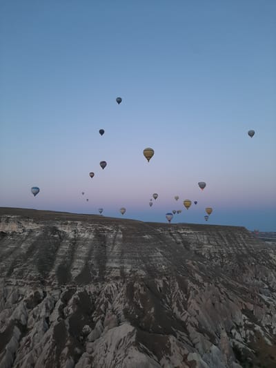 The image shows a panoramic view of hot air balloons ascending over the rocky, eroded landscape of Cappadocia, near Avanos, Turkey. The sky transitions from a pale blue at the top to a soft pink and purple near the horizon, indicating either sunrise or sunset. Numerous hot air balloons of various colors and sizes are scattered across the sky, some appearing higher than others. The foreground features a dramatic, layered geological formation characterized by steep cliffs and ravines, with sparse vegetation. The overall scene depicts a serene and breathtaking aerial spectacle.
