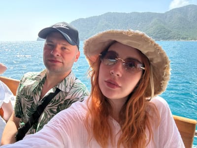 Two individuals are positioned closely, facing the camera in an upside-down orientation. The environment features blue water and a hazy, mountainous backdrop, suggestive of an outdoor setting, possibly a boat excursion.
The person on the left has red hair, wears a neutral-colored sun hat and rimless sunglasses, and is dressed in a white top. The person on the right wears a dark baseball cap and a light-colored, tropical-patterned shirt.
The geographical location, Kumluca, Turkey is inferred from available information about the imagery, it is unclear what is their action or interaction in the image.