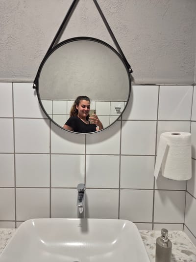 A young woman with brown hair pulled back in a ponytail is taking a mirror selfie in a bathroom. She is wearing a black t-shirt and is smiling. The bathroom has white tiled walls with grey grout. A round mirror hangs on the wall above a white sink. To the right of the mirror, a roll of toilet paper is mounted on the wall. The sink has a chrome faucet and a textured countertop. A soap dispenser is visible on the countertop to the right of the sink. The overall setting appears to be a restroom. This scene is located in Kazichene, Bulgaria.