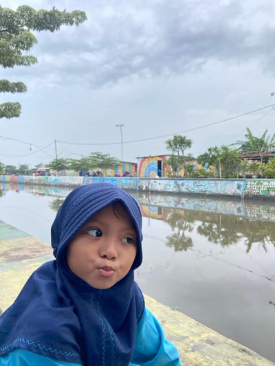 A young child, appearing to be Southeast Asian, is captured in a mid-shot, positioned in the foreground to the left. The child wears a dark blue, pleated hijab with light blue stitching and a light blue garment beneath. Their eyes are directed towards the upper right, and the mouth is pursed. The child is situated on a yellowish-grey concrete embankment with faint discolorations.

Immediately adjacent to the child is a placid, brownish-grey body of water, likely a canal or river, which occupies the central horizontal plane of the image. The water reflects the overcast sky and elements from the opposite bank.

The far bank features a concrete wall adorned with vibrant, multicolored murals and graffiti. Behind this decorated wall, several small, simple structures are visible, some painted with bright stripes and a large rainbow motif. Sparse green vegetation, including trees and shrubs, dots the far bank. In the distance, power lines traverse the scene, supported by poles, and a street light fixture is discernible. Faint human figures and what appear to be parked motorcycles or vehicles are visible on the distant bank. The sky above is overcast with uniform grey cloud cover.

The scene depicts a developed waterway area within the city of Kalitengah, Indonesia.