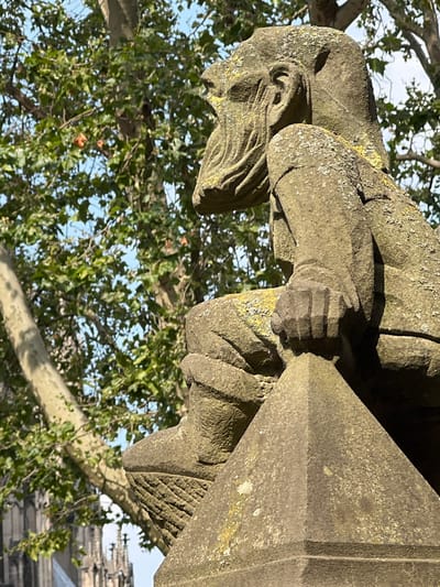 This is a close-up, eye-level outdoor photograph of a stone gargoyle or grotesque sculpture, weathered and partially covered in lichen, positioned in front of a tree with lush green leaves and a bright blue sky. The sculpture is depicted from the chest up, with its head turned to the left, showing a profile view of its face, including an ear and a long, flowing beard. Its left arm is bent, with its hand resting on its lap or a surface. The sculpture is seated on a pointed stone base. In the background, blurred by distance, are Gothic-style architectural elements, consistent with structures found in Cologne, Germany, such as those of the Cologne Cathedral. The lighting suggests a sunny day.