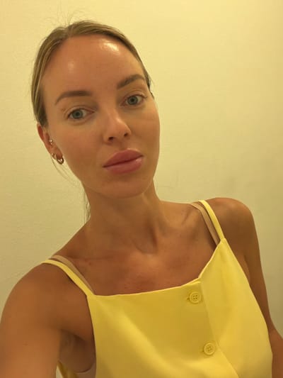 A medium shot shows a woman from the chest up, looking towards the camera. She has fair skin, blue eyes, and blonde hair pulled back. She is wearing a yellow camisole with thin straps and two buttons down the front. She is also wearing small hoop earrings. The background is a plain, pale yellow wall. The image appears to be a selfie. There is no information to suggest the location is in Finestrat, Spain.