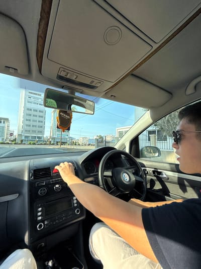 The image shows the interior of a car, viewed from the passenger's seat, with a young man driving. The car is on a street in Larnaca, Cyprus, evidenced by the architecture of the buildings visible through the windshield, which are typical of the city. The driver is wearing sunglasses and a dark blue polo shirt. His right hand is on the steering wheel, and his left hand is near the hazard light button. The dashboard features a standard car stereo and air vents. Outside the car, a modern multi-story building with many windows is prominent, along with other city buildings and streetlights. An "areon" air freshener hangs from the rearview mirror. The sky is clear and blue, suggesting a sunny day.