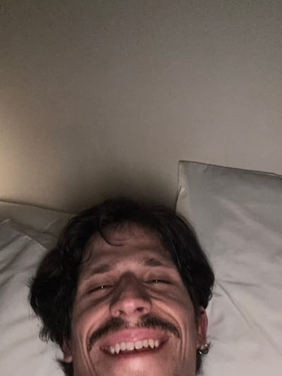 A person is lying on their back in bed with a white pillow visible behind their head. The person has dark hair and a mustache, and they are smiling with their mouth open, revealing their teeth. An earring is visible in their ear. The background includes a plain, light-colored wall. The location is unknown.