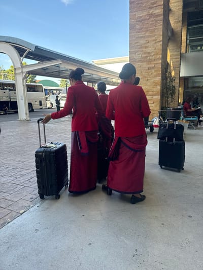 Three women in matching red uniforms stand with their luggage on a paved outdoor area. They are likely at an airport or transit station. One woman is pulling a black suitcase. Behind them, a bus is visible. In the background, there is a building with a stone facade and what appears to be an elevated train track structure. Additional luggage is stacked nearby. The scene appears to be captured on a bright, sunny day.