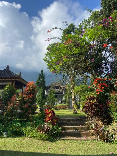 This is an outdoor, eye-level, vertical photograph taken in Sukasada, Indonesia. The scene depicts a lush garden with a variety of plants and flowers, including trees with pink and red blossoms. A stone staircase is visible on the right side of the frame, leading upwards. In the background, there is a building with dark windows and a dark, cloudy sky overhead. A traditional Balinese-style pavilion with an ornate roof is visible in the lower part of the image, partially obscured by foliage. The lighting suggests it might be late afternoon or early morning, with shadows cast by the trees and the pavilion. The overall atmosphere is serene and tropical, characteristic of a garden setting in Indonesia. There is no visible text in the image.