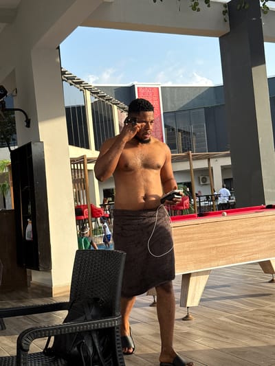 The image depicts a shirtless man in a dark towel standing next to a pool table on a patio in Rubuchi, Nigeria. He is wearing sandals and appears to be talking on a cell phone. The patio has wooden floors and is surrounded by a building with glass windows and a railing. There are people visible in the background near a pool. A black wicker chair is in the foreground.