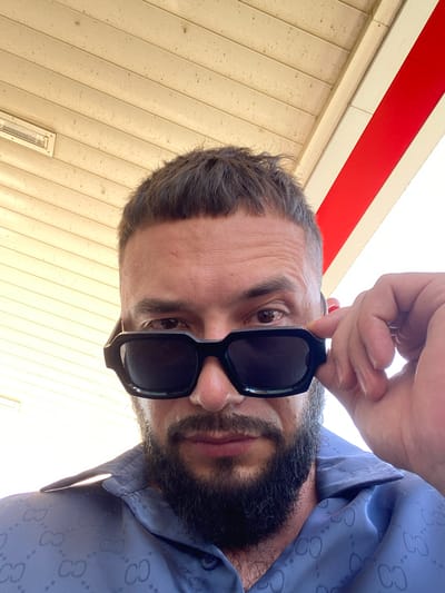 The image shows a close-up, low-angle selfie of a man. He is wearing black, rectangular sunglasses and a blue collared shirt with a repeating 'GC' pattern. His hand is near his sunglasses, adjusting them. The background appears to be an outdoor structure with a light-colored, slatted ceiling and a red stripe. The scene suggests a casual setting, possibly a street or a covered outdoor area in Korkmaskala, Russia.