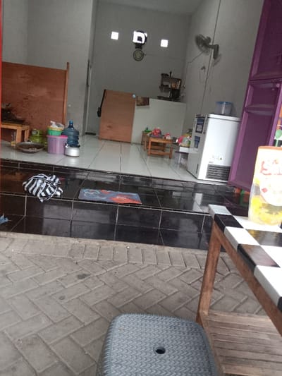 The image shows the exterior of a small storefront, likely a food vendor, located at coordinates -6.945, 110.221 in Kecamatan Brangsong, Indonesia. A small, rectangular, white refrigerator stands to the right of the doorway, with a purple door behind it. There is a wooden table with a black and white checkerboard top in the foreground to the right. There is a black and white striped tote bag lying on the steps leading up to the storefront, and a black and orange doormat on the step closest to the interior. There is a clock hanging on the wall inside. Two small tables with chairs sit to the right of the doorway, along with a variety of containers, including a large blue water jug. The floor is tiled in white and black tiles. The environment appears to be a densely populated urban area. The image is taken from a low angle, with the camera pointed upwards towards the storefront. There is a low black stool in the lower right corner of the image. There are no people visible in the image.