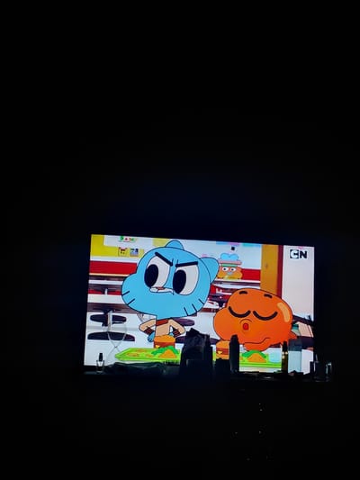 The image shows a television screen displaying a scene from the animated series "The Amazing World of Gumball". The characters Gumball Watterson (blue, cat-like) and Darwin Watterson (orange, fish-like) are visible. Gumball is looking to the left with a stern expression, while Darwin is looking down with closed eyes, seemingly eating a hamburger. In the background, there are shelves with various items and a "CN" logo, indicating Cartoon Network. The television is placed on a surface that appears to be a table or shelf, in front of a dark, out-of-focus background. Some faint lights are visible below the screen. The scene is not located in the city of Malorad, Bulgaria.