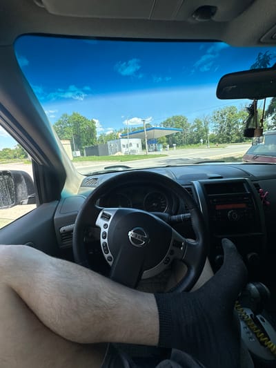 The image is a first-person perspective from inside a car, looking towards the driver's side. The driver's legs, one wearing a black sock and the other bare, are visible. The steering wheel, a Nissan, is in the foreground. Beyond the steering wheel, the dashboard of the car is visible, including the radio and air vents. The windshield shows a bright blue sky with scattered clouds, suggesting a clear day. Outside the car, a gas station with a blue canopy is visible, along with trees and a road. The image appears to be taken during the daytime, likely mid-day given the brightness of the sky. There is no clear indication of the specific location within Shatava, Ukraine, beyond the presence of a gas station and surrounding vegetation. The overall impression is a casual scene of someone in a car on a sunny day.
