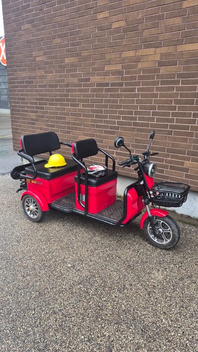 A bright red electric scooter with two seats is parked on a gravel surface in front of a brown brick wall. A yellow hard hat is placed on the front seat, and a white and red helmet rests on the rear seat. The scooter has a basket mounted on the front for storage. The scooter appears to be a three-wheeled model with a single wheel at the front and two wheels at the rear. The environment suggests an outdoor urban setting, possibly near a building with a brick facade. The image is taken from a slightly elevated angle, looking down at the scooter and its surroundings.