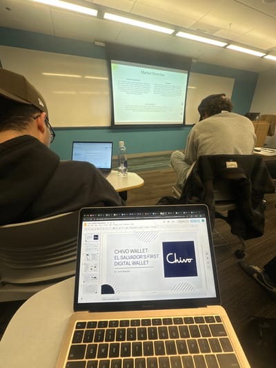 A classroom scene features a student engrossed in a laptop displaying a presentation slide titled "CHIVO WALLET: EL SALVADOR'S FIRST DIGITAL WALLET" by Sofia Maestre. Simultaneously, a projector screen in the background shows "Market Overview." Other students are present in the room. A water bottle rests on a desk beside a second laptop. The scene is illuminated by overhead lighting.