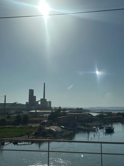 This is a vertically oriented, landscape photograph of an industrial complex situated alongside a body of water. The sun is brightly shining, creating lens flare and a bright halo effect. The sky is clear and appears hazy, suggesting a bright, possibly warm day. On the left side of the frame, a dark wire runs diagonally from the bottom left corner upwards.
The industrial complex consists of several buildings with visible smokestacks, pipes, and various structures. There are also signs of disuse or abandonment in some parts of the complex. The water in the foreground is calm, with reflections of the sky and structures. There are a few small boats moored or docked in the water. The background shows rolling hills or mountains under a hazy sky. A fence or railing runs across the right side of the image, suggesting the viewpoint is from a raised structure or a balcony overlooking the scene.
No people are visible in the image. The overall mood is somewhat stark and industrial, juxtaposed with the natural element of the water and the bright, hazy sunlight. The time of day is likely daytime, given the strong sunlight. The location is identified as Bages, France. There is no visible text in the image.