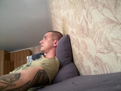 A man with a shaved head and tattoos on his arm is lying on a bed, looking up and to the left. He is wearing a green t-shirt. The bed is in a room with a patterned wallpaper and a wooden closet in the background. The image appears to be taken indoors in a residential setting.