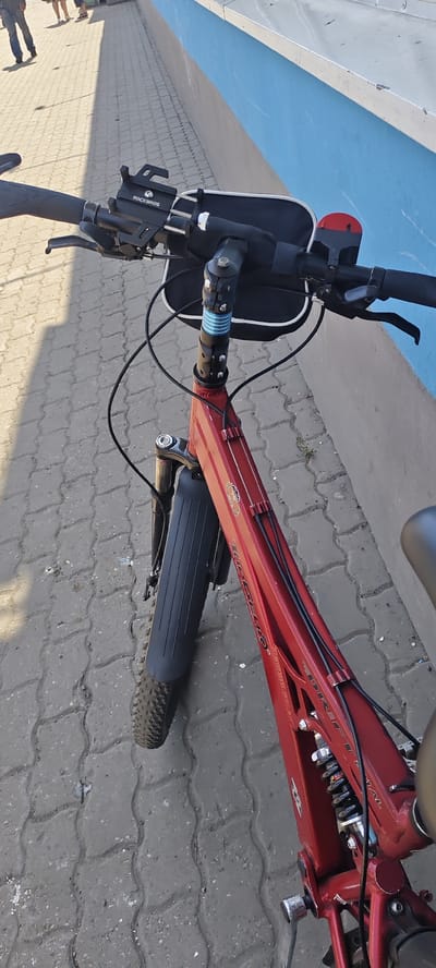 A red electric bicycle is parked on a cobblestone sidewalk next to a blue and gray painted wall. The bicycle has a phone mount on the handlebars with the "ROCKBROS" logo visible. A black bag is attached to the handlebars, and a red bell is also present. The bicycle's frame has "TREK" and "FULL" written on it. The front wheel has a black fender. In the background, the legs of two people are visible, walking away from the camera. The scene appears to be outdoors in an urban environment.