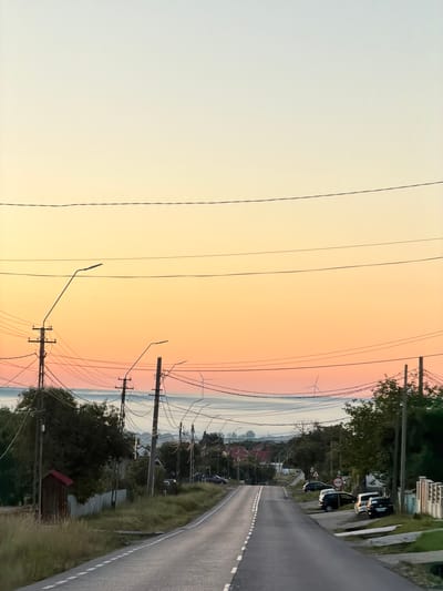 This is a forward-facing, eye-level, medium shot of a paved road in the city of Pașcani, Romania, during what appears to be sunrise or sunset. The sky exhibits a gradient of colors, transitioning from pale yellow at the top to orange and pink hues closer to the horizon. Several utility poles with numerous wires are visible along the sides of the road, as well as streetlights. On the left side of the road, there is a small wooden structure with a red roof, surrounded by tall grass and foliage. Further down the road on the right, several parked cars are visible, including a dark-colored sedan, a black SUV, a white hatchback, and another dark-colored sedan. A sign with red and white circles is posted near the parked cars. Trees and houses are scattered throughout the background, with distant hills or low mountains partially obscured by mist or haze. In the far distance, a wind turbine is faintly visible against the colorful sky. The road itself has a white dashed line marking the center. The overall scene depicts a rural or semi-rural setting on the outskirts of a town.