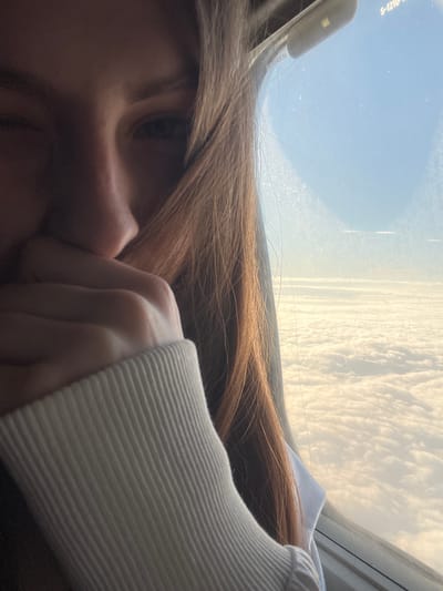 The image shows a close-up of a young person's face, with their hand covering their mouth, as they look out of an airplane window. The person has long, light brown hair that partially obscures their face. Outside the window, a vast expanse of white clouds is visible under a clear blue sky. The scene appears to be taking place on a flight, with the light and angle suggesting it could be during sunrise or sunset. The location is identified as Stolbishche, Russia.