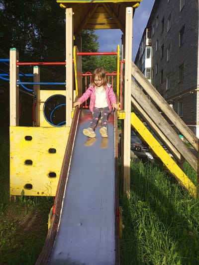 A young child, identified as a girl with light brown hair and eyeglasses, is captured in motion on a metal slide at a playground in Berezniki, Russia. She is dressed in a pink jacket over a white top, patterned leggings, and light-colored sneakers, with her hands resting on the slide's worn, reddish-brown and yellow edges as she descends. The slide is part of a larger, multi-component playground structure constructed from yellow painted wood and various colored metal bars (red, blue), exhibiting visible signs of wear and chipped paint. To the left of the structure, dense green tree foliage is visible against a partially cloudy blue sky. To the right, a multi-story residential building, constructed of light grey brick with numerous windows and enclosed balconies, stands prominently. A grey vehicle, possibly a Dacia/Renault Duster, with a partially visible "MA" on its license plate, is parked beneath the residential building, partially obscured by the playground structure and overgrown green grass. The ground beneath and around the playground is covered in tall green grass and some dirt patches. The scene is illuminated by direct sunlight, creating distinct shadows.