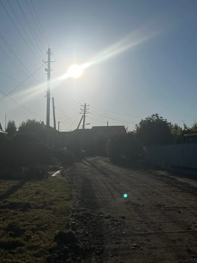 This image depicts a rural or suburban street scene in Tikhoretsk, Russia, bathed in bright sunlight, causing significant lens flare. The foreground features a dirt road or path with scattered pebbles and some sparse vegetation on the left side. In the middle ground, silhouetted against the sun, are the roofs of houses and dense foliage. Numerous power lines crisscross the sky, supported by wooden utility poles, with one prominent pole in the left-center of the frame. A distinct bright green circular light artifact is visible on the dirt road in the lower right quadrant of the image, likely a lens artifact from the bright sunlight. The overall impression is that of a sunny, possibly late afternoon, rural landscape.