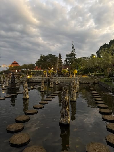 The image depicts a serene outdoor scene at what appears to be a temple or garden complex in Abang, Indonesia. The main subjects are the architectural elements and the natural surroundings. In the foreground, a body of water is crossed by a path of octagonal stepping stones, each decorated with a circular motif. Stone sculptures, possibly of deities or mythical figures, stand partially submerged in the water or along the edges of the water features, adding to the cultural and spiritual atmosphere.

Beyond the water, a traditional Balinese building with a tiered roof is visible, along with other temple-like structures. The landscape is lush with mature trees and manicured greenery. Strategically placed lights illuminate certain areas, suggesting it is dusk or early evening. The sky is filled with dramatic, cloudy formations, with patches of lighter, possibly sunlit, clouds creating a contrast.

The overall mood is peaceful and contemplative, enhanced by the soft lighting and the still water reflecting the structures and the sky. There are no visible people in the frame, focusing the viewer's attention on the tranquil beauty of the location. The time of day is clearly not midday, given the low light and dramatic sky. The presence of ornate stonework and distinct architectural styles strongly suggests a location within Indonesia, consistent with the provided location context of Abang. No legible text is present in the image.