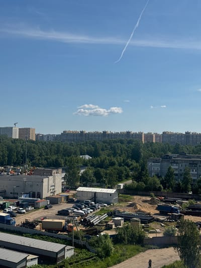 This is a high-angle, full-shot photograph of an industrial and residential area in Yanino-1, Russia. The sky is bright blue with some wispy clouds and a contrail from an aircraft. In the background, there are rows of tall apartment buildings, some under construction, surrounded by a dense forest. Closer to the foreground, there is an industrial yard with various buildings, vehicles, and materials. This includes several light-colored industrial buildings, a white modular building, a blue semi-trailer truck, a large beige container on a trailer, piles of sand, and stacks of large black pipes. Several cars are parked in the yard. In the lower right corner, two figures are seen riding a scooter on a dirt path. The overall scene depicts a blend of urban development, industrial activity, and natural landscape.