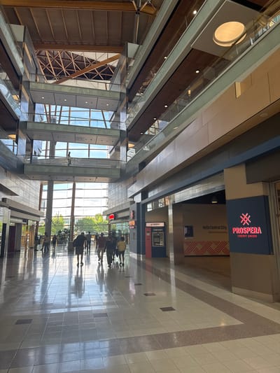 The image shows the interior of a modern building, likely a shopping mall or office complex, with multiple levels and an atrium. Natural light streams in from large windows, illuminating a polished tile floor that reflects the surrounding architecture and the people walking through. Various architectural elements are visible, including exposed wooden beams in the ceiling, glass railings on the upper levels, and polished metal accents.
To the right of the image, a prominent sign for "PROSPERA CREDIT UNION" is visible, with the text "Hello Central City" and "Welcome to local banking" also displayed. Adjacent to the sign is an ATM machine. Several individuals are walking in the main corridor of the building. Their attire suggests a casual setting.
This scene is located at the Central City Shopping Centre in Surrey, British Columbia, Canada.