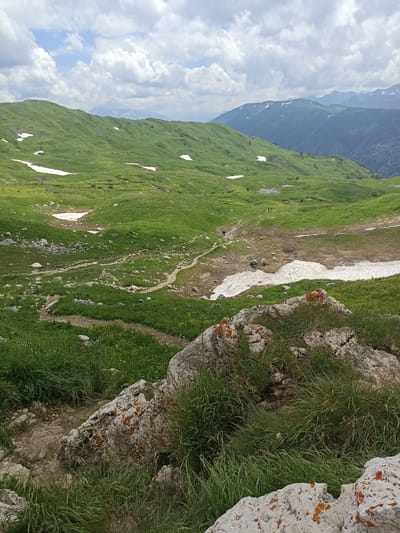 The image depicts a vast, mountainous landscape with rolling green hills and patches of snow. The sky is filled with white, fluffy clouds against a light blue backdrop. In the foreground, large rocks covered in orange lichen and green grass are visible. A dirt path winds through the terrain, leading towards a shallow body of water or wet ground. In the distance, several figures, appearing small due to the distance, are walking along the path. Further in the background, more mountain ranges are visible, some with snow-capped peaks. The overall environment suggests a natural, outdoor setting, likely a hiking trail in a mountainous region. The location provided, Chigursana, Russia, indicates a high-altitude area with mountainous terrain.