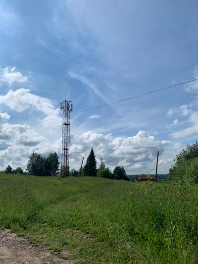 The image depicts a cellular tower standing tall in a grassy field under a partly cloudy sky. The tower, predominantly red and grey metal lattice, is equipped with multiple antennas. A dirt path leads towards the tower through the lush green grass. In the background, a line of trees separates the field from a small cluster of buildings, one of which has a rusty brown roof. Power lines crisscross the scene, supported by wooden poles. The overall environment suggests a rural or semi-rural setting in the city of Barsai, Russia. No people are visible in the frame.