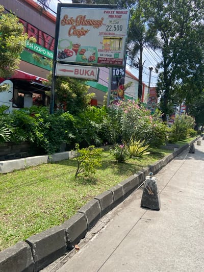 A street scene in Margaasih, Indonesia (latitude: -6.958, longitude: 107.561), showing a sidewalk bordered by a concrete curb and a grassy area. Two large rocks with a blue and white object are placed in the sidewalk and are connected with string. A sign on a pole reads "Sate Maranggi Cianjur" and displays a price of 22,500 Rupiah. A sign that reads "BUKA" is also visible. A building is in the background with green foliage. A red truck with an advertisement for a store can be seen behind the sign.