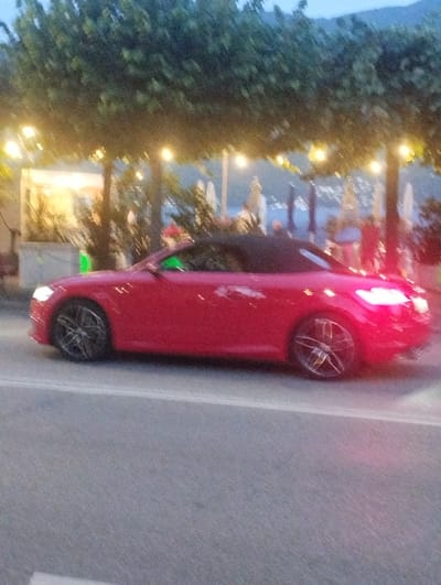 A red convertible car is captured in a side profile, driving on a paved road. The car's top is down, revealing a black interior. The car's headlights are illuminated, casting a bright white light. The car is positioned on the right side of the frame, with its front facing towards the right. Behind the car, a line of trees with decorative lights strung between them is visible. Beyond the trees, there appears to be a body of water, possibly a lake, with some structures and umbrellas on the opposite shore. The sky is a dusky blue, suggesting it is either early morning or late evening. The general setting appears to be a scenic lakeside location, consistent with the city of Lugano, Switzerland, which is known for its beautiful lake and surrounding mountains. The image is slightly blurry due to motion, but the key elements of the car and the environment are discernible.