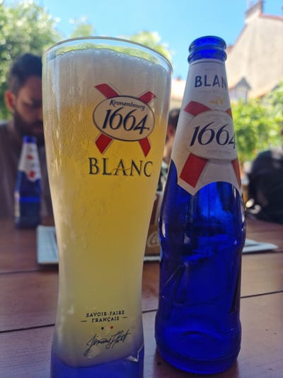 A glass of Kronenbourg 1664 Blanc beer and a blue bottle of the same brand are on a wooden table outdoors. The glass is tall and has the Kronenbourg 1664 Blanc logo, featuring an oval with the number 1664 and a red and yellow emblem. The bottle also has a label with "BLANC" and "1664 BIERE BLANCHE" printed on it. The beer in the glass is pale yellow and frothy. In the background, a person is visible, out of focus, and there are trees and a building, suggesting an outdoor seating area in a city. The scene appears to be set in Cetinje, Montenegro, as indicated by other images from the same location.