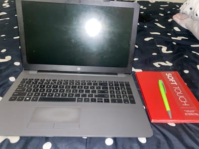 An HP laptop is positioned on a bed with a black and white patterned sheet. The screen is dark, and the keyboard is visible. Adjacent to the laptop is a red "Soft Touch" notebook. A green pen lies on top of the notebook. The scene suggests a casual work or study setup.
