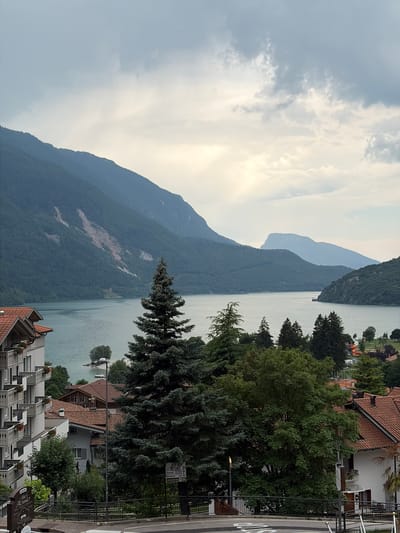 The image depicts a scenic view of Lake Molveno in Molveno, Italy. In the foreground, a multi-story white building with balconies is visible, adjacent to several residential buildings with red-tiled roofs. A dense forest of evergreen and deciduous trees occupies the middle ground. The lake itself is a tranquil body of water reflecting the overcast sky. In the background, a series of imposing, verdant mountains rise dramatically, with one slope showing evidence of a landslide. The sky is cloudy with patches of light breaking through. The overall atmosphere is serene and natural, characteristic of a mountain lakeside town. There are no people or animals clearly visible in the image.