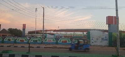 The image shows a street in Abakaliki, Nigeria, at sunset. The sky is a light pink and orange color, and there are numerous power lines running overhead. A blue tricycle is parked on the right side of the road. In the background is a large building with a green and blue facade. The building appears to be a market or a bus station. There are a few people walking around, and the building is mostly empty. There are green plants growing in the front of the building. The scene is calm and quiet, with the only sound being the hum of traffic in the distance. The coordinates provided place the location of the image near the Abakaliki Central Market.